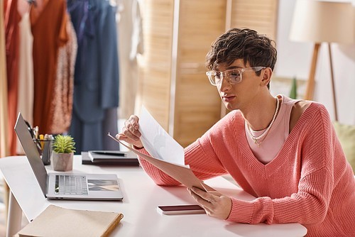 curly androgynous freelancer in pink sweater and eyeglasses working remotely near devices on desk