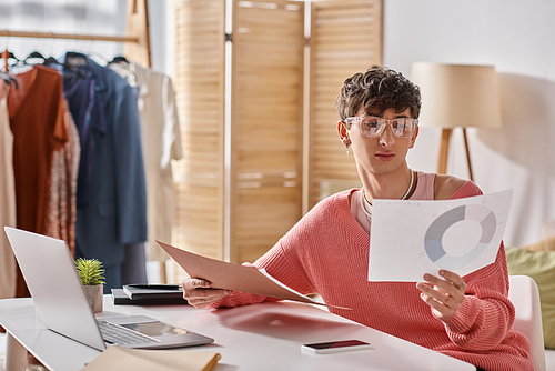 curly androgynous freelancer in pink sweater and eyeglasses working remotely near devices on desk