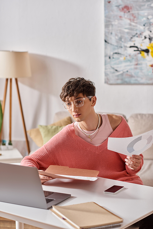 curly androgynous freelancer in pink sweater and eyeglasses working remotely near devices on desk