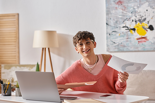 happy androgynous blogger in pink sweater and eyeglasses working remotely near devices on desk