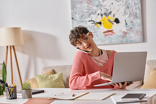 cheerful androgynous freelancer in pink sweater and eyeglasses using laptop and smartphone at home