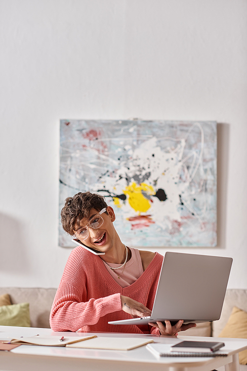 cheerful androgynous freelancer in pink sweater and eyeglasses using laptop and smartphone at home