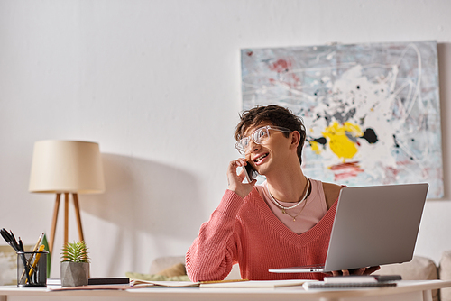 pleased androgynous freelancer in pink sweater and eyeglasses using laptop and smartphone at home