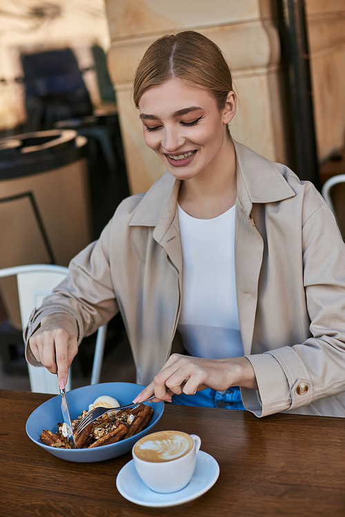 happy young woman in trench coat eating her belgian waffles with ice cream next to cup of coffee