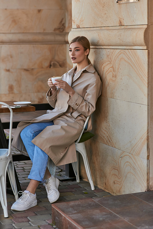 beautiful young woman in stylish trench coat sitting and holding cup of coffee inside of modern cafe