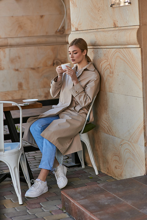 attractive young woman in stylish trench coat sitting and holding cup of coffee inside of cafe