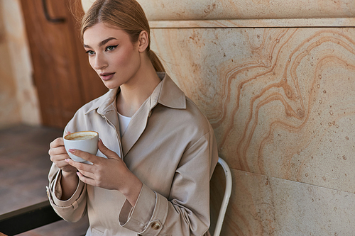 blonde young woman in stylish trench coat sitting and holding cup of coffee inside of modern cafe