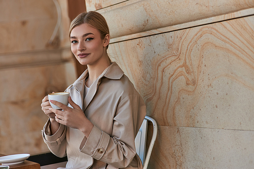 happy young woman in stylish trench coat sitting and holding cup of coffee inside of modern cafe