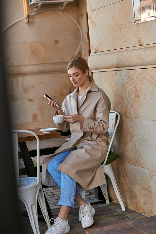 attractive young woman in stylish trench coat using smartphone and holding cup of coffee in cafe