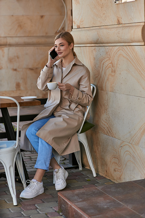happy young woman in stylish trench coat talking on smartphone and holding cup of coffee in cafe