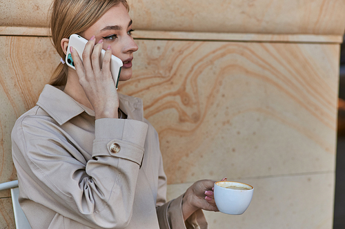 young woman in stylish trench coat talking on smartphone and holding cup of coffee in cafe