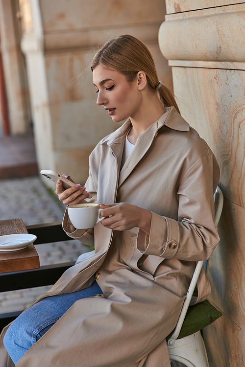attractive young woman in stylish trench coat using smartphone near cup of coffee on table in cafe