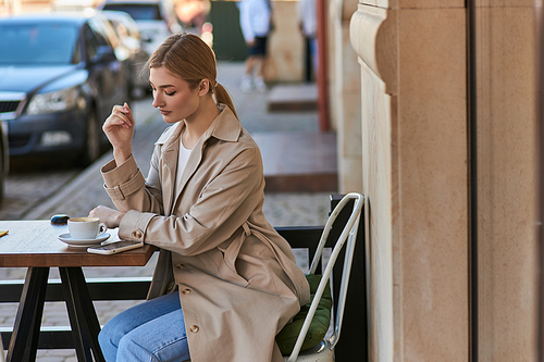 blonde woman in stylish trench coat using smartphone near cup of coffee on table in cafe