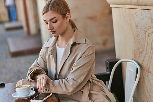 blonde young woman in stylish trench coat using smartphone near cup of coffee on table in cafe