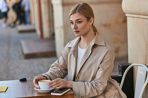 blonde young woman in trench coat sitting near cup of coffee and smartphone on table in cafe