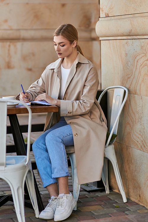 blonde young woman in trench coat taking notes in notebook near cup of coffee in cafe, planning day