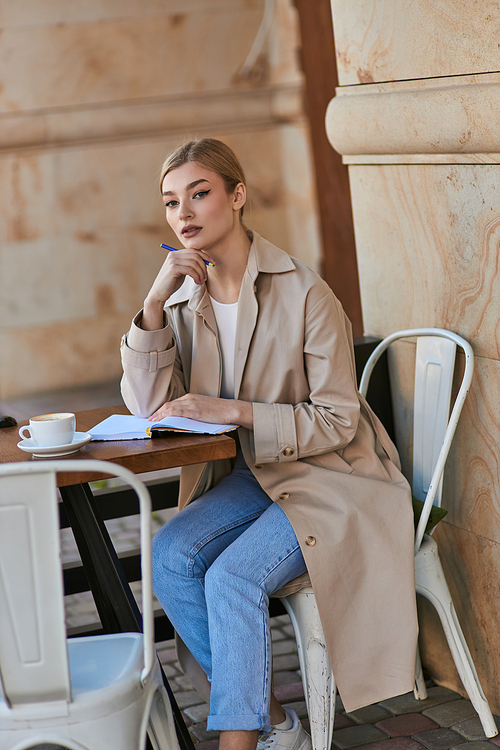 pensive blonde woman in trench coat sitting at table and holding pen near chin, writing diary
