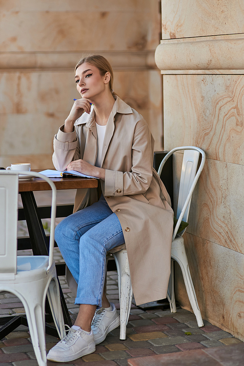 thoughtful blonde woman in trench coat sitting at table and holding pen near chin, writing diary