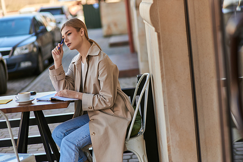 thoughtful woman in trench coat sitting at table with cup of coffee and holding pen, writing diary