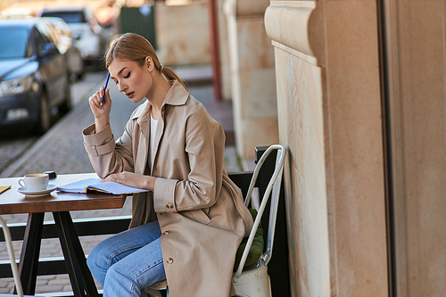 pensive woman in trench coat sitting at table with cup of coffee and holding pen, writing diary