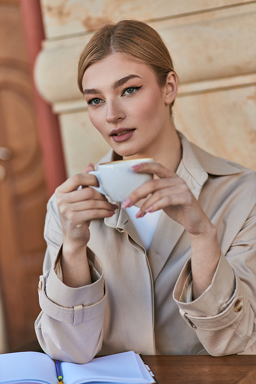 elegant blonde woman in stylish trench coat holding cup of coffee while sitting in cafe, beverage
