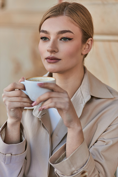 pretty blonde woman in stylish trench coat holding cup of coffee while sitting in cafe, beverage