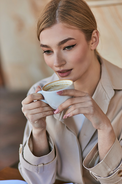 elegant blonde woman in stylish trench coat holding cup with coffee while sitting in cafe, beverage