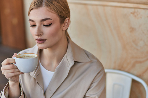elegant blonde woman in stylish trench coat holding cup of coffee while sitting in cafe, cappuccino