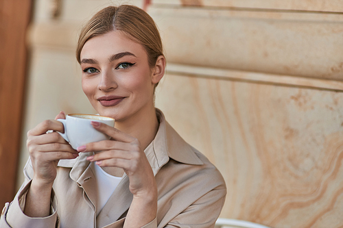 cheerful blonde woman in stylish trench coat holding cup of coffee while sitting in cafe, beverage