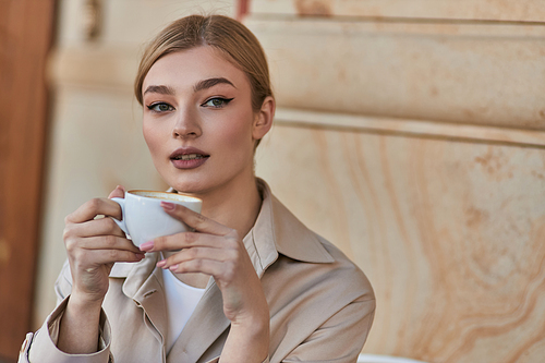 elegant young woman in stylish trench coat holding cup of coffee while sitting in cafe, beverage
