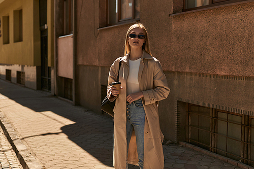 elegant woman in stylish trench coat holding takeaway coffee while standing on street in sunny day