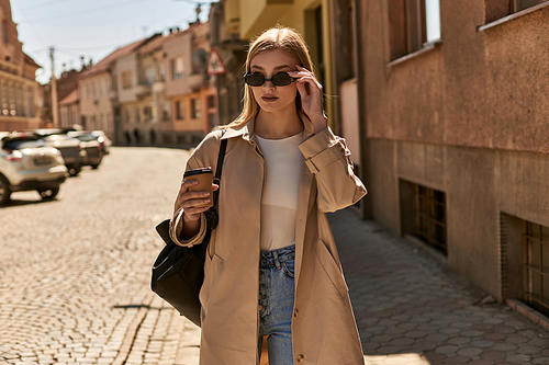 young blonde woman in trench coat holding takeaway coffee while standing on street in sunny day