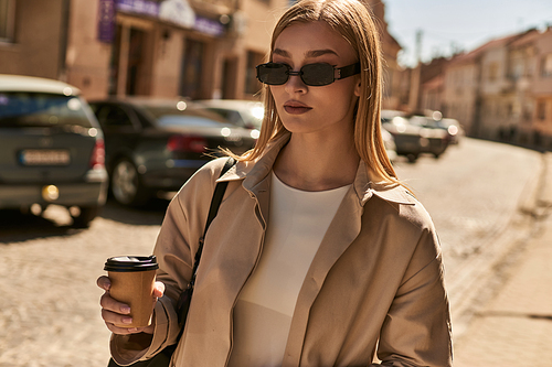 blonde woman in stylish trench coat holding coffee to go while standing on street in sunny day