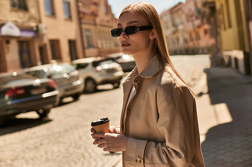 blonde woman in stylish trench coat holding coffee to go while walking on street in sunny day