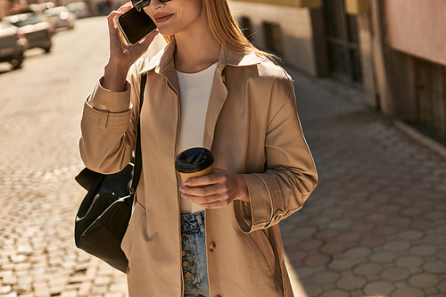 cropped view of young woman in trench coat talking on smartphone and holding coffee to go on street