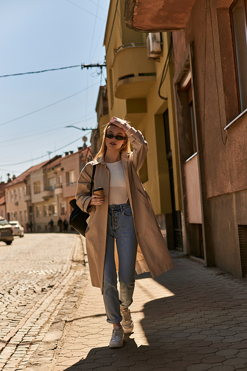 stylish young woman in trench coat and sunglasses holding coffee to go and walking on street