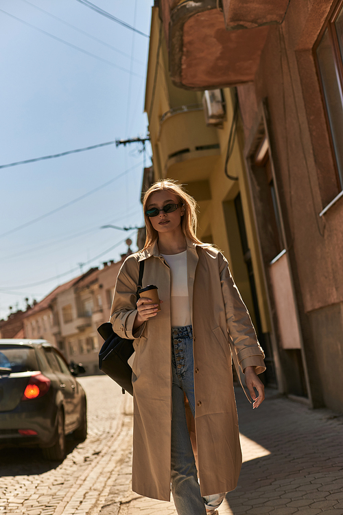 blonde young woman in trench coat and sunglasses holding coffee to go and walking on street