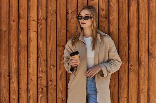 blonde young woman in trench coat and sunglasses holding coffee to go and standing near wooden fence