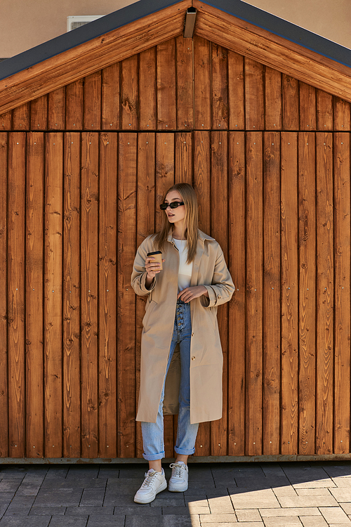 young woman in stylish trench coat and sunglasses holding coffee to go and standing near fence