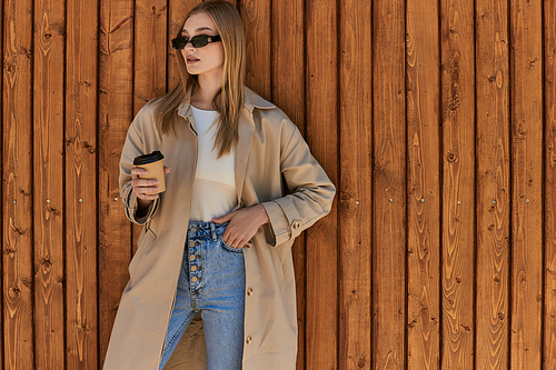 blonde woman in stylish trench coat and sunglasses holding coffee to go and standing near fence