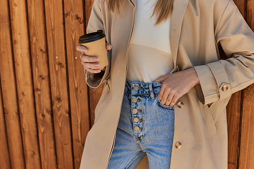 cropped woman in stylish trench coat and sunglasses holding coffee to go and standing near fence