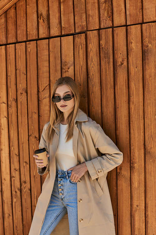 blonde woman in trench coat and sunglasses holding coffee to go and standing with hand in pocket