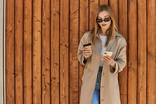 blonde woman in trench coat and sunglasses holding coffee to go and using smartphone near fence