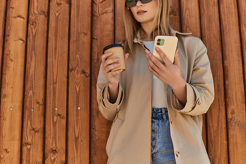 cropped woman in trench coat and sunglasses holding coffee to go and using smartphone near fence