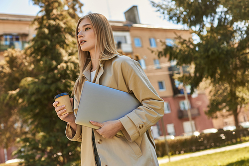 stylish blonde woman in trench coat walking with laptop and takeaway coffee, freelancer in city