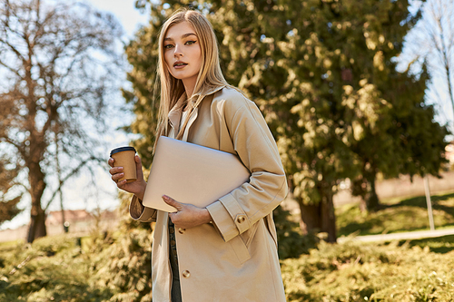 young blonde woman in trench coat walking with laptop and takeaway coffee, freelancer in city