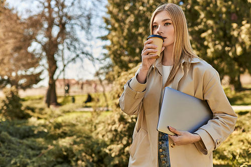 young blonde woman in trench coat holding laptop and drinking takeaway coffee, freelancer in city