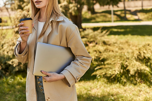cropped young woman in trench coat holding laptop and drinking takeaway coffee, freelancer in city
