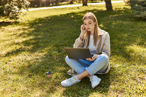 young blonde woman in earphones and trench coat sitting on grass near paper cup and using laptop