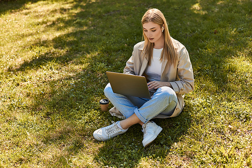 blonde woman in earphones and trench coat sitting on grass near paper cup and using laptop